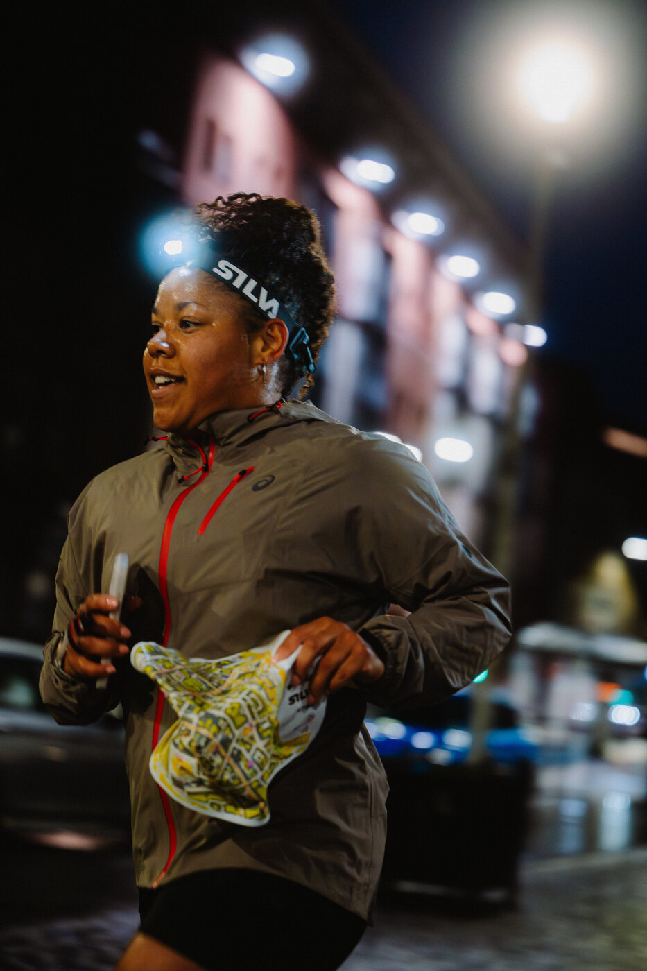 An orienteer in a grey jacket holding a map and wearing a headtorch at night